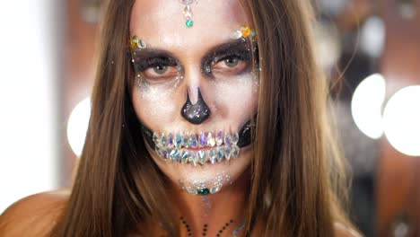 Chica-joven-asustadiza-con-creativa-halloween-de-pintura-de-guerra-en-el-camerino.-Retrato-de-glamour-cráneo-con-pedrería-y-lentejuelas.-Maquillaje-profesional-para-la-celebración.-Cámara-lenta