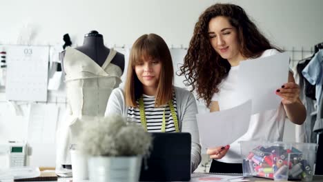 Besitzer-von-Schneiderei-Studio-arbeitet-mit-Laptop-Arbeitsplatz,-während-ihre-Designer-mit-Skizzen-zu-ihr-kommt.-Frauen-sind-Bildschirm-und-Zeichnungen-betrachten-und-diskutieren-Kleidung.