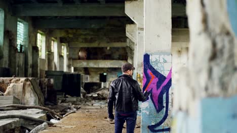 Male-graffiti-painter-is-creating-abstract-image-with-spray-paint-inside-abandoned-empty-building.-Old-column-is-in-foreground,-dirty-walls-and-windows-in-background.