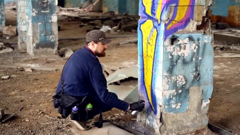 Graffiti-artist-bearded-guy-is-painting-on-pillar-in-abandoned-building-with-aerosol-paint-spray.-Empty-industrial-building-with-dirty-walls-and-floor-is-in-background.