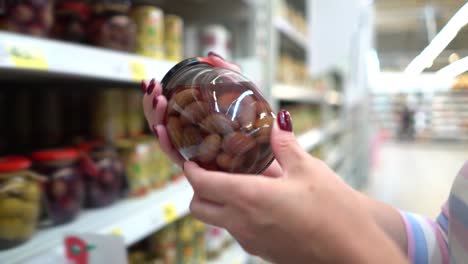 Manos-de-mujer-caucásica-de-Closeup-cerca-de-estantes-de-la-tienda-eligiendo-escabeche-aceitunas-negras-abarrotes-mercado