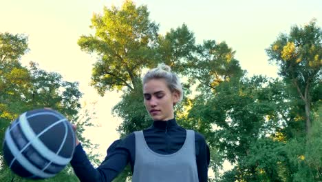 Charming-female-basketball-player-walking-and-playing-with-ball,-park-with-blue-sky-above-in-background,-low-angle