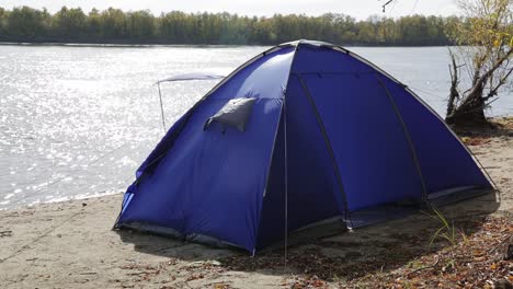 A-lonely-blue-tent-blown-up-by-a-strong-wind-on-the-Bank-of-an-autumn-river