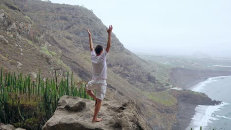 Un-hombre-de-pie-en-el-borde-de-un-acantilado-con-vistas-al-océano-levanta-sus-manos-e-inhala-el-aire-del-mar-durante-el-yoga