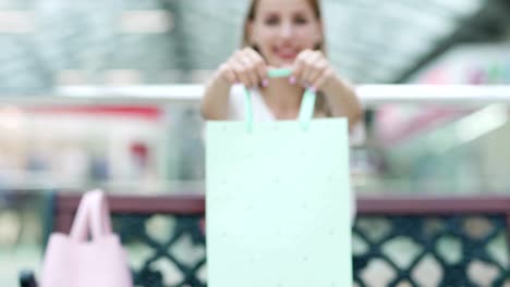 Fade-in-of-satisfied-young-woman-sitting-on-bench-in-shopping-mall-and-showing-mint-shopping-bag-with-purchased-item