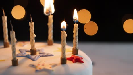 Close-up-of-four-candles-being-lit-with-a-match,-amongst-unlit-candles-on-a-white,-decorated-birthday-cake,-bokeh-lights-in-the-background