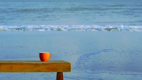 Orange-cup-of-coffee-on-brown-wooden-table-on-beach-with-surf-of-blue-sea-in-background.