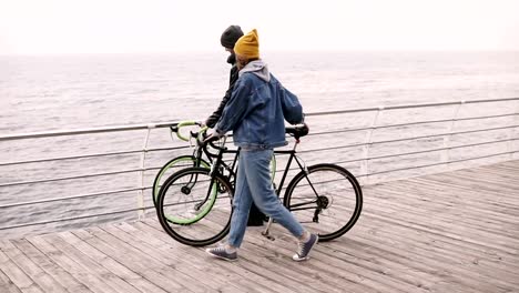 Pareja-encantadora-hipster-deja-sus-motos,-apoyado-en-la-barra-caminando-juntos-abrazando-cerca-del-mar-en-día-de-otoño.-Caminando-por-cubierta-de-madera-durante-el-día