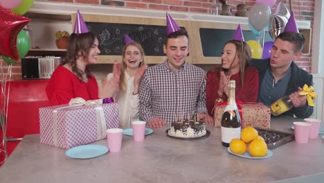 Handsome-man-blowing-birthday-candles-while-close-friends-throwing-a-surprise-party,-slowmotion-in-cozy-modern-apartment