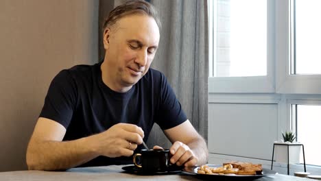 Un-hombre-de-mediana-edad-en-la-mañana-tomando-café-y-comiendo-galletas-hechas-en-casa.-Él-se-sienta-en-casa-en-la-cocina-y-él-está-de-buen-humor.