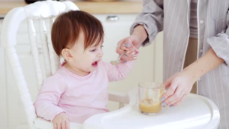 Madre-irreconocible-ayudando-a-su-hija-bebé-a-comer-puré-de-frutas-con-cuchara-en-la-trona