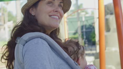 Mujer-sonriendo-mientras-sostiene-al-bebé-en-el-portaequipajes-en-un-patio-de-recreo