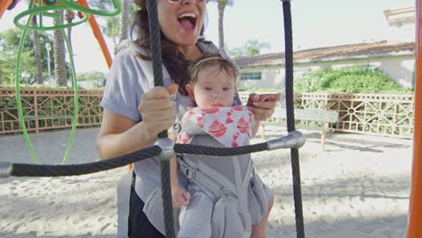 Mujer-con-bebé-en-el-portador-divirtiéndose-en-el-patio-de-recreo