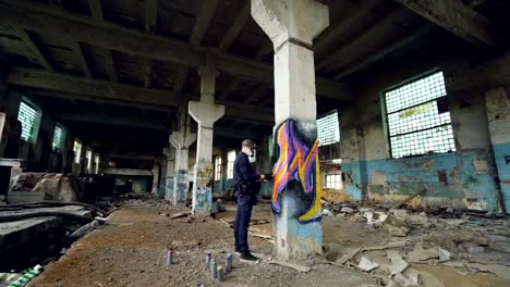 Graffiti-artist-in-protective-mask-is-painting-on-high-column-in-abandoned-industrial-building.-Creative-people,-modern-wall-art-and-protective-equipment-concept.