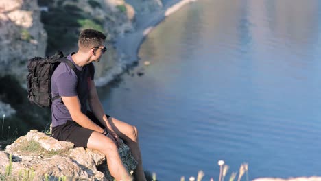 Side-view-handsome-European-young-male-traveler-sitting-on-cliff-near-clean-pond-medium-long-shot