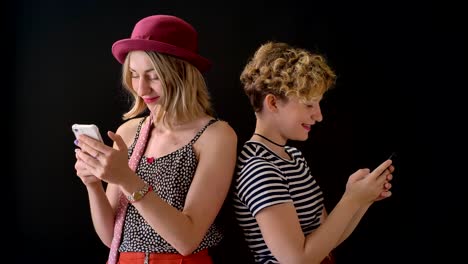 Two-happy-young-women-texting-on-phone-and-smiling,-standing-isolated-on-black-background