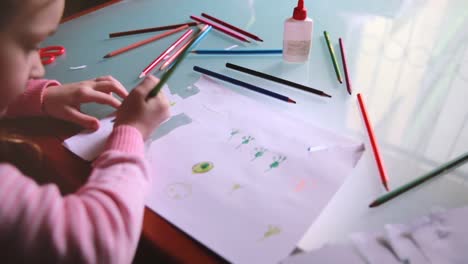 Top-view-camera-sliding-left-on-little-Caucasian-girl-child-drawing-on-paper-with-various-color-pencils-at-a-table