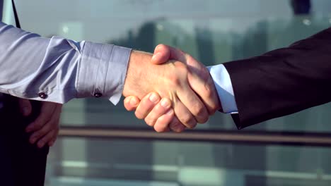 Close-up-of-successful-businessmen-greeting-each-other-near-office-building.-Two-young-colleagues-meeting-and-shaking-hands-in-urban-environment.-Handshake-of-business-partners-outdoor.-Side-view