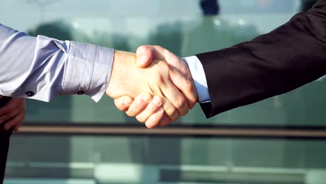 Two-confident-businessmen-shaking-hands-after-successful-deal-near-office-building.-Young-colleagues-congratulating-each-other-in-urban-environment.-Handshake-of-business-partners.-Slow-motion