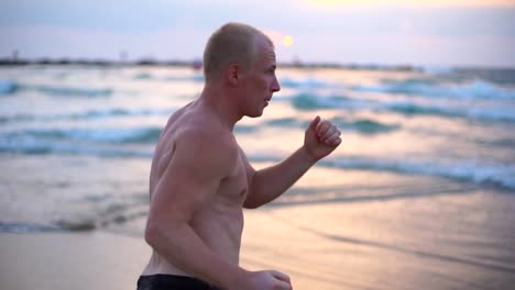 Joven-musculoso-hombre-practicando-ejercicios-de-boxeo-en-la-playa-del-mar.-Deportista-masculino-es-practica-defensa-personal-sola-cerca-de-la-orilla-del-mar.-Entrenamiento-de-artes-marciales-al-aire-libre-al-atardecer.-Lenta-de-cerca