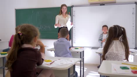 Grupo-escuela-de-los-niños-sentados-y-escuchando-classmate-junto-a-la-pizarra-con-el-maestro-durante-la-clase-en-aula-de-espalda