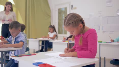 kleines-Schulmädchen-sitzt-hinter-der-Schulbank-während-Zeichnung-Lektion-in-helle-Klassenzimmer-auf-Hintergrund-der-Mitschüler-und-eine-junge-Lehrerin