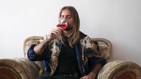 Happy-young-man-sitting-on-a-comfortable-armchair-with-cushions-wearing-a-jacket-inside-his-home-holding-a-glass-of-delicious-red-wine-tasting-and-swirling