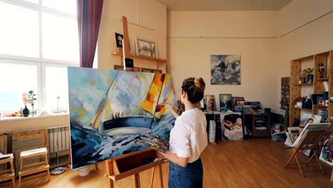 Pan-shot-of-female-painter-depicting-marine-landscape-on-canvas-using-oil-paints,-brush-and-palette-working-in-workshop-alone.-Visual-arts-and-youth-concept.