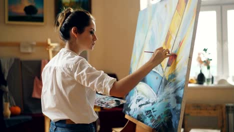 Professional-artist-young-woman-is-painting-seascape-with-acrylic-paints-finishing-marine-landscape-ship-and-sea-waves-working-alone-in-cozy-studio.-People-and-work-concept.