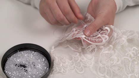 A-close-up-of-woman's-hands-sewing-white-beads-lying-in-the-round-lid-in-front-of-her-to-ornamented-fabric