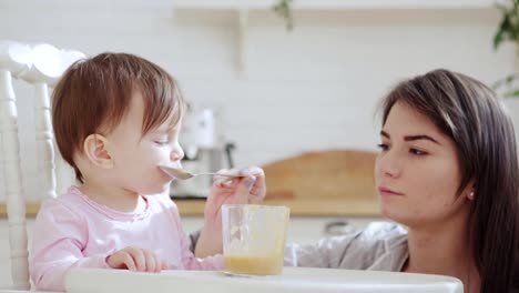 Close-up-side-view-of-young-mother-feeding-her-baby-daughter-vegetable-puree-with-spoon,-girl-trying-to-feed-herself