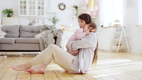 Young-mother-lying-on-hardwood-floor-in-domestic-room-embracing-her-cute-baby-daughter