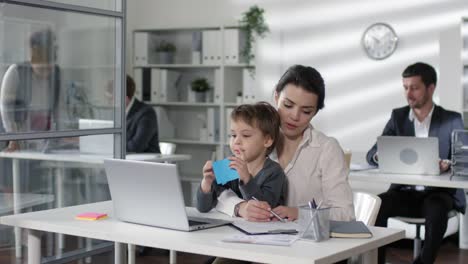 Geschäftsfrau-arbeitet-am-Schreibtisch-mit-Kind-auf-Knien