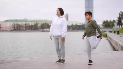 Asian-Mother-and-Son-Doing-Stretching-Exercise-on-Urban-Riverside-Sidewalk