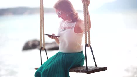 Young-happy-woman-wearing-sunglasses-uses-mobile-phone-on-a-swing-on-a-tropical-beach-and-splashes-waves-beautiful-breaks-by-stones-in-the-sea.-1920x1080