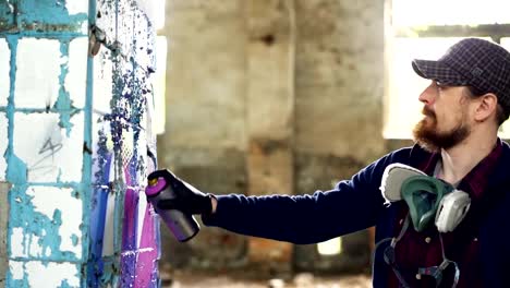 Attractive-bearded-guy-is-holding-aerosol-paint-and-drawing-graffiti-on-pillar-inside-spacious-abandoned-house.-Creative-people,-empty-buildings-and-modern-art-concept.