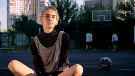 Wunderschöne-blonde-Frau-auf-Basketballplatz-lächelnd-in-die-Kamera,-Männer-im-Hintergrund-spielen,-weibliche-Spieler