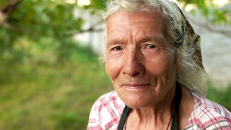 Portrait-of-an-elderly-woman-in-a-kerchief.-The-wind-wavers-her-gray-hair,-she-looks-into-the-camera-and-blinks.-Grandmother-in-the-garden-near-the-house.-Close-up