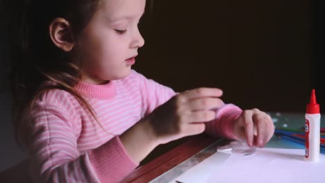 Close-up-shot-of-beautiful-little-preschool-European-girl-child-in-pink-sweater-sitting-by-the-table-gluing-paper-shapes