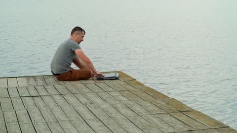 a-man-is-approaching-to-the-end-of-a-masonry-and-opening-a-laptop-on-the-background-of-lake-in-the-summer