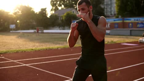 Concentrated,-muscular-man-boxer-doing-boxing-exercise,-warming-up-while-standing-on-stadium-outdoors-in-summer-sun-rays.-Shadow-fight