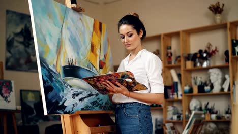 Skillful-female-painter-is-working-in-studio-alone-painting-picture-on-easel-using-oil-paints,-palette-and-brush-enjoying-her-occupation.-People-and-work-concept.