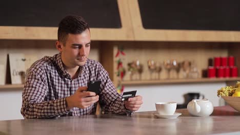 Handsome-man-making-purchase-online,-typing-credit-card-details-on-black-smartphone-in-the-modern-kitchen