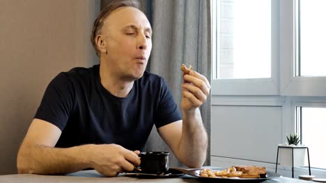 The-man-has-breakfast-in-the-morning-at-home-in-the-kitchen,-and-he-is-in-a-good-mood.-He-drinks-coffee-and-eating-homemade-cookies.