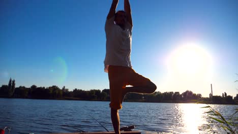 Young-guy-practicing-yoga-position-on-the-edge-of-wooden-jetty-at-lake-on-summer-day.-Sporty-man-doing-exercise-near-lake-with-sunlight-at-background.-Healthy-active-lifestyle.-Slow-motion-Close-up