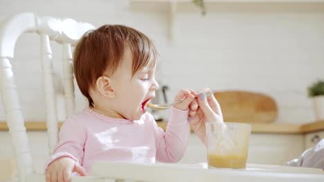 Vista-lateral-de-niña-linda-sentada-en-silla-alta-y-comiendo-con-puré-de-verduras-de-cuchara-ayudada-por-su-madre-irreconocible