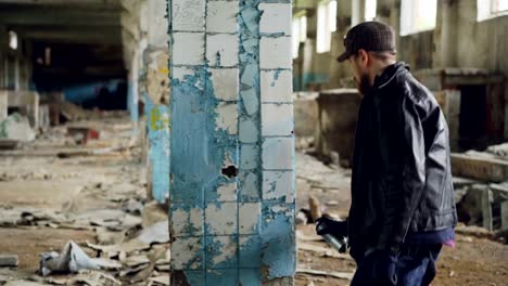 Graffiti-artist-bearded-guy-is-painting-on-pillar-in-abandoned-building-with-bright-aerosol-paint-spray.-Pan-shot-of-empty-industrial-building-with-dirty-walls-and-broken-windows.