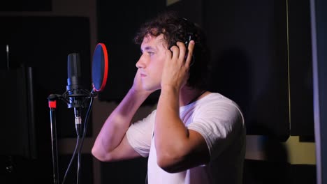 Young-handsome-singer-man-puts-on-headphones-in-the-studio.-Recording-new-melody-or-album.-Male-vocal-artist-with-curly-hair-preparing-for-working.-Slow-motion