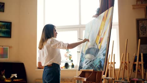 Pretty-girl-painter-is-mixing-paints-on-palette-then-painting-seascape-on-canvas-creating-beautiful-picture.-Nice-modern-studio-with-tools-is-visible.