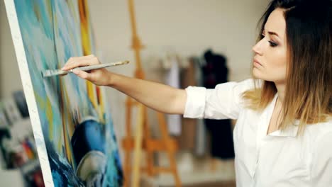 Pretty-young-lady-is-busy-painting-picture-in-workshop-using-bright-paint-and-stained-palette.-Girl-is-wearing-casual-clothing-and-is-concentrated-on-her-work.